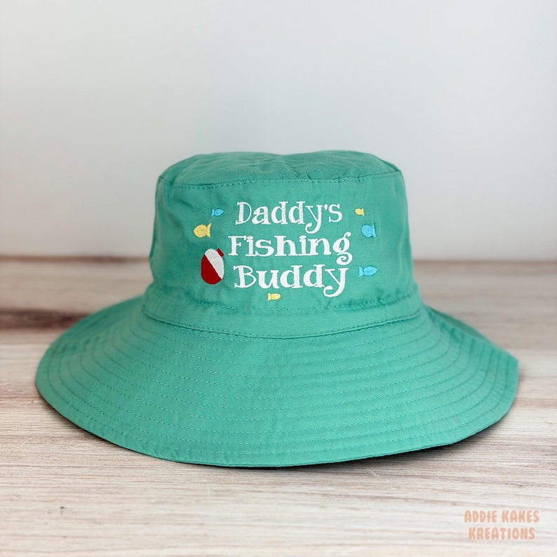 May include: A teal green fishing hat with a wide brim. The hat features the embroidered text "Daddy's Fishing Buddy" in white, along with small fish and a beach ball graphic. The hat is designed for sun protection.