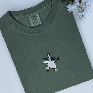 May include: Olive green t-shirt with a crew neck and the words "SILLY GOOSE" above a cartoon goose graphic. The shirt has a tag that reads "COMFORT COLORS". The shirt is folded on a white surface.