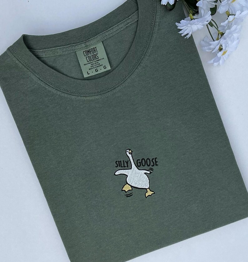 May include: Olive green t-shirt with a crew neck and the words "SILLY GOOSE" above a cartoon goose graphic. The shirt has a tag that reads "COMFORT COLORS". The shirt is folded on a white surface.