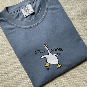 May include: A slate blue crew neck t-shirt featuring an embroidered graphic of a white goose with yellow feet and the black text SILLY GOOSE. The interior neck label displays the brand name Comfort Colors and the size XL-TG-EG.