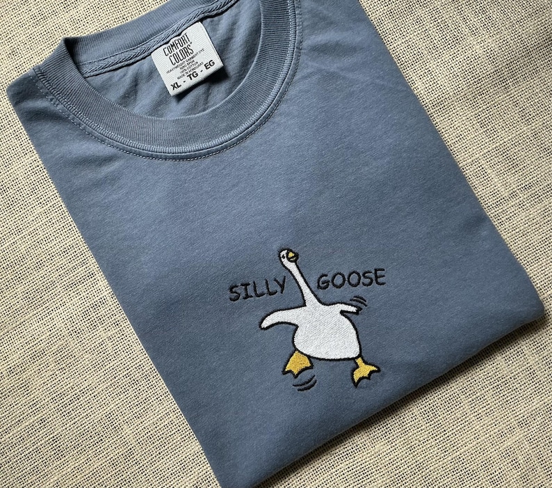 May include: A slate blue crew neck t-shirt featuring an embroidered graphic of a white goose with yellow feet and the black text SILLY GOOSE. The interior neck label displays the brand name Comfort Colors and the size XL-TG-EG.