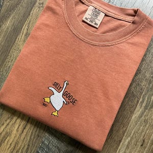 May include: A coral-coloured t-shirt with a crew neck and short sleeves. The shirt features an embroidered design of a white goose with yellow feet and the words "Silly Goose". The tag reads "Comfort Colors".