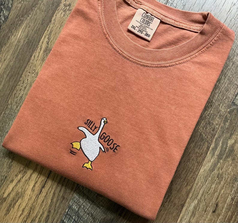 May include: A coral-coloured t-shirt with a crew neck and short sleeves. The shirt features an embroidered design of a white goose with yellow feet and the words "Silly Goose". The tag reads "Comfort Colors".