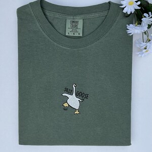 May include: An olive green t-shirt featuring the embroidered phrase "Silly Goose" and a goose graphic. The shirt is folded and has a Comfort Colors label. White flowers are in the upper right corner.