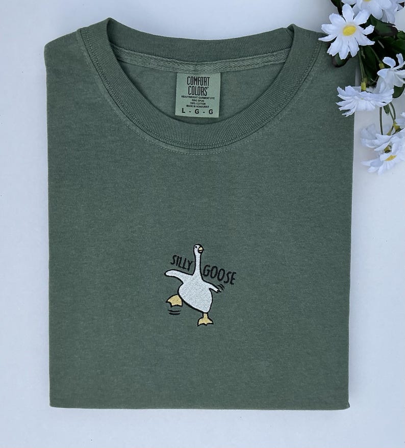 May include: An olive green t-shirt featuring the embroidered phrase "Silly Goose" and a goose graphic. The shirt is folded and has a Comfort Colors label. White flowers are in the upper right corner.