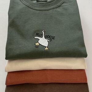 May include: Four t-shirts in a stack, in shades of olive green, cream, rust, and brown. The top shirt is olive green and features the embroidered text "Silly Goose" and a cartoon goose.