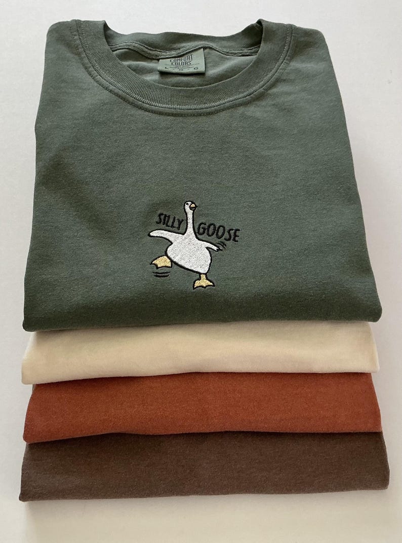 May include: Four t-shirts in a stack, in shades of olive green, cream, rust, and brown. The top shirt is olive green and features the embroidered text "Silly Goose" and a cartoon goose.