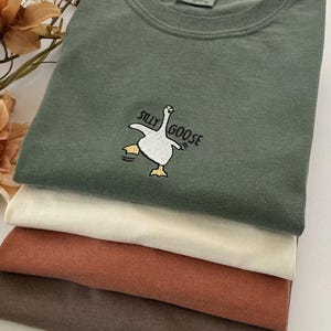 May include: A stack of folded t-shirts in shades of green, cream, rust, and brown. The top shirt is green with a "Silly Goose" embroidered design. The design features a white goose with yellow feet and the text "Silly Goose" in black.