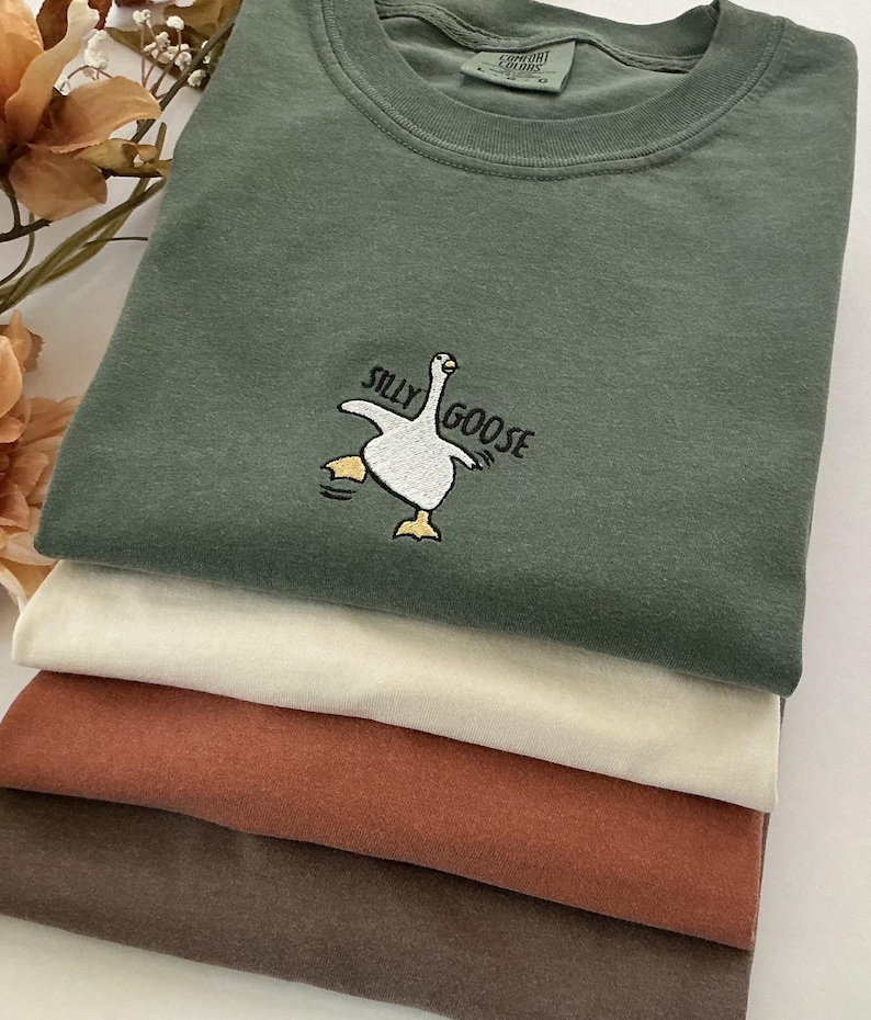 May include: A stack of folded t-shirts in shades of green, cream, rust, and brown. The top shirt is green with a "Silly Goose" embroidered design. The design features a white goose with yellow feet and the text "Silly Goose" in black.