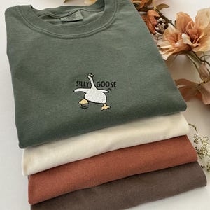 May include: A pile of folded t-shirts in shades of olive green, cream, rust, and brown. The top shirt, olive green, features an embroidered "Silly Goose" design. The shirts are stacked, suggesting a casual clothing theme.