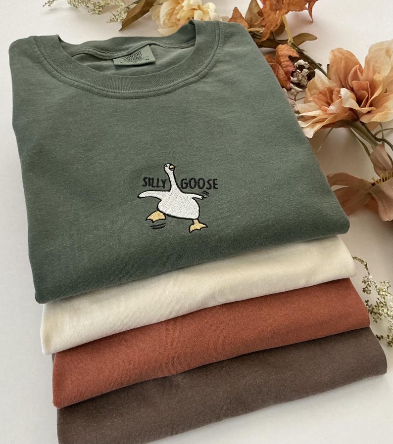 May include: A pile of folded t-shirts in shades of olive green, cream, rust, and brown. The top shirt, olive green, features an embroidered "Silly Goose" design. The shirts are stacked, suggesting a casual clothing theme.