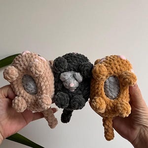 May include: Three handmade crochet animal toys in a row. The toys are in shades of tan, gray, and orange. Each has a gray center, pink accents, and a small tail. The toys are held up against a white background.