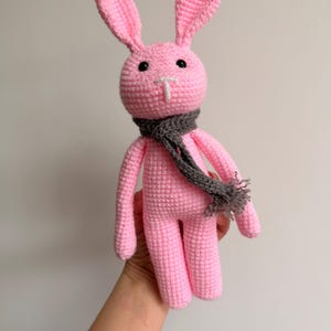 Puede incluir: Un conejo de ganchillo hecho a mano en color rosa claro, con orejas largas y erguidas, ojos de seguridad de plástico negro y una nariz bordada en blanco. El conejo lleva una bufanda de punto gris oscuro con flecos en el cuello.