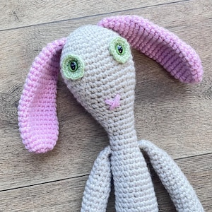 May include: A crocheted bunny toy with pink ears and green eyes. The bunny is made of a soft, beige yarn and has a pink nose.