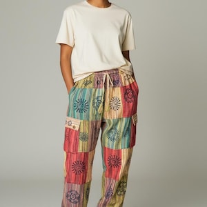May include: A person wearing a cream-colored t-shirt and colorful patchwork pants. The pants feature a variety of patterns and colors, including red, yellow, green, and blue. The pants have pockets and a drawstring waist. The person is also wearing beige sneakers.