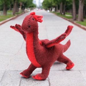May include: A red felt dragon toy with a long neck and a long tail. The dragon is standing on a gray sidewalk.