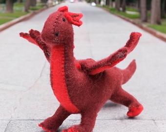 May include: A red felt dragon toy with a long neck and a long tail. The dragon is standing on a gray sidewalk.