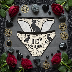 May include: Black and white underwear featuring a black cat graphic and the text "Hexy and I Know It". The underwear is surrounded by black stones, pentagrams, and red roses.