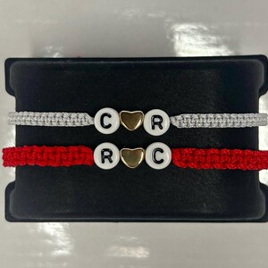 Puede incluir: Dos pulseras de la amistad con hilo rojo y blanco, cada una con un dije de corazón dorado y cuentas blancas con letras negras que deletrean "C R" y "R C".