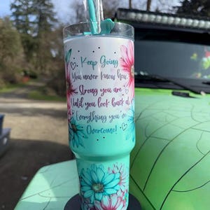 May include: A teal tumbler with a floral design and the text "Keep Going, You never know how. Strong you are until you look back at everything you've overcome!" The tumbler has a clear lid and a teal straw.