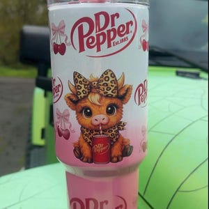 Pode incluir: Um copo Dr. Pepper rosa e branco com um desenho de vaca de desenho animado. O copo apresenta o logotipo da Dr. Pepper e cerejas. Inclui uma palhinha e tampa rosa. A vaca está usando um laço e um lenço com estampa de leopardo.
