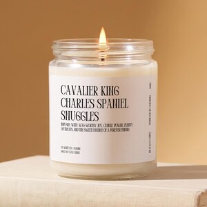 May include: A clear glass jar candle with a white label featuring black text that reads Cavalier King Charles Spaniel Snuggles, infused with wag-worthy joy, cuddle power, plenty of treats, and the sweet essence of a forever friend.