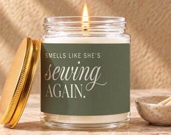 May include: A clear glass jar candle with a lit wick and a gold-colored metal lid. The label is olive green with white text that reads: SMELLS LIKE SHE'S sewing AGAIN. The candle is filled with white wax and emits a warm glow from the flame.