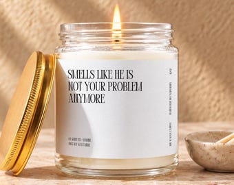 May include: A clear glass jar candle with a gold lid and a white label featuring the black text: SMELLS LIKE HE IS NOT YOUR PROBLEM ANYMORE. The candle contains white wax and has a lit wick. Additional text on the label includes 100% SOY WAX CANDLE.