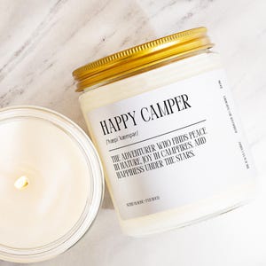 A white candle labeled "Happy Camper" with a gold lid, placed next to a partially visible candle on a marble surface.