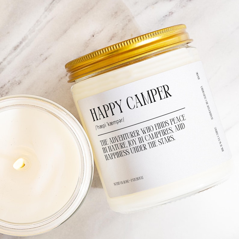 A white candle labeled "Happy Camper" with a gold lid, placed next to a partially visible candle on a marble surface.