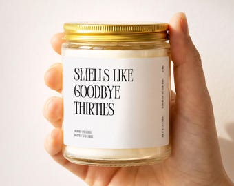 May include: A clear glass candle jar with a gold metal lid containing white wax. The white label features black text that reads: SMELLS LIKE GOODBYE THIRTIES, ROSE + PATCHOULI, 100% SOY WAX CANDLE, HANDMADE IN CALIFORNIA, 8 OZ WAX CANDLE.