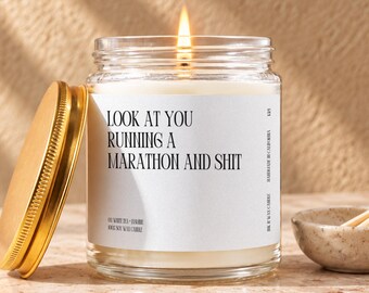 May include: A lit soy wax candle in a clear glass jar with a gold metal lid. The white label features black text reading: LOOK AT YOU RUNNING A MARATHON AND SHIT, OH WHITE TEA + JASMINE, 100% SOY WAX CANDLE, HANDMADE IN CALIFORNIA, 8 OZ WAX CANDLE.