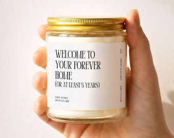 May include: A clear glass candle jar with a gold metal lid containing white wax. The white label features black text that reads: WELCOME TO YOUR FOREVER HOME (OR AT LEAST 5 YEARS), along with smaller text indicating rose and patchouli scent.