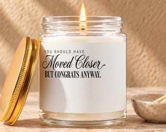 May include: A clear glass candle jar with a gold metal lid and a white label featuring the black text: YOU SHOULD HAVE Moved Closer BUT CONGRATS ANYWAY. The candle contains white wax and has a lit wick centered in the middle of the jar.