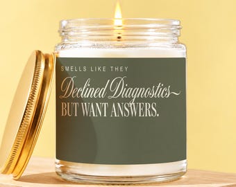May include: A clear glass candle jar with a lit wick and a gold lid. The label is olive green with white text that reads: SMELLS LIKE THEY Declined Diagnostics BUT WANT ANSWERS. The candle is filled with white wax and emits a warm, steady flame.