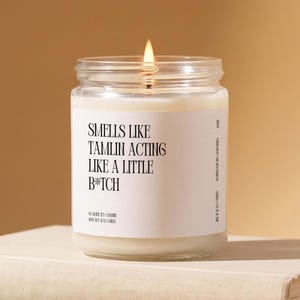 May include: A clear glass jar candle with a white label featuring black text that reads: SMELLS LIKE TAMLIN ACTING LIKE A LITTLE B*TCH. The candle contains off-white wax and has a single lit wick centered at the top, emitting a warm glow.