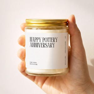 May include: A clear glass candle jar with a gold metal lid containing white wax. The white label features black text that reads HAPPY POTTERY ANNIVERSARY, along with smaller text indicating rose, patchouli, and soy wax candle ingredients.