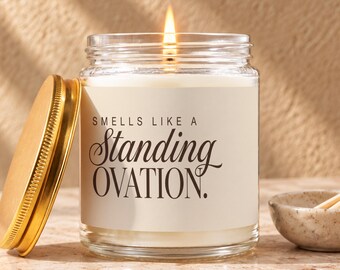 May include: A clear glass jar candle with a lit wick and a gold-colored metal lid resting beside it. The white label features the black text: SMELLS LIKE A Standing OVATION. The candle contains white wax and is filled to the top of the glass jar.