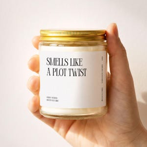 May include: A clear glass candle jar with a gold metal lid containing white wax. The white label features black text reading "SMELLS LIKE A PLOT TWIST" with smaller text below stating "OI ROSE + PATCHOULI" and "100% SOY WAX CANDLE" and "HANDMADE IN CALIFORNIA".