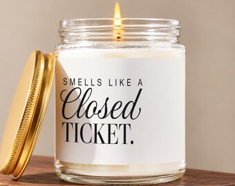May include: A clear glass candle jar with a white label featuring the black text "SMELLS LIKE A Closed TICKET." The candle has a lit wick and is accompanied by a separate, round, metallic gold lid placed to the left of the glass jar.