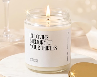 May include: A clear glass jar candle with a white label featuring the black text IN LOVING MEMORY OF YOUR THIRTIES. The candle has a lit wick with a bright flame. A gold-colored metal lid sits beside the jar on a white circular base.