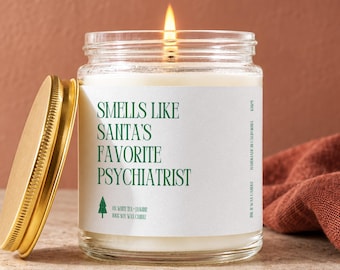 May include: A clear glass jar candle with a lit wick and a gold-colored metal lid. The white label features green text that reads: SMELLS LIKE SANTA'S FAVORITE PSYCHIATRIST, along with a small green tree icon and the text 100% SOY WAX CANDLE.