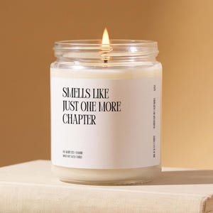 May include: A clear glass jar candle with a lit wick and white wax. The white label features black text reading: SMELLS LIKE JUST ONE MORE CHAPTER, TEA WHITE TEA + JASMINE, 100% SOY WAX CANDLE, HANDMADE IN CALIFORNIA, 8OZ.