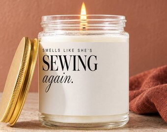 May include: A clear glass jar candle with a lit wick and a gold-colored metal lid placed to the side. The white label features the black text: "SMELLS LIKE SHE'S SEWING again." The candle contains white wax and emits a warm, steady flame.