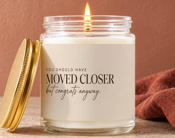 May include: A clear glass candle jar with a lit wick and a gold-colored metal lid. The label features the text: YOU SHOULD HAVE MOVED CLOSER but congrats anyway. The candle contains white wax and is presented in a simple, cylindrical glass container.