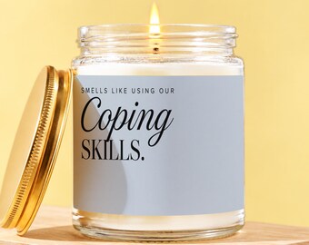 May include: A clear glass candle jar with a lit wick and a gold-colored metal lid placed to the side. The label is light blue with black text that reads: SMELLS LIKE USING OUR Coping SKILLS.