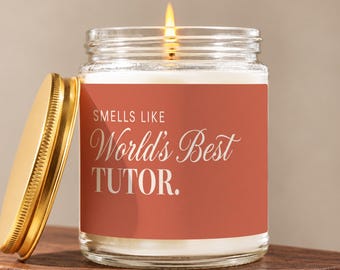 May include: A clear glass candle jar with a lit wick and a gold-colored metal lid placed to the side. The jar features a terracotta-colored label with white text that reads: SMELLS LIKE World's Best TUTOR. The candle wax inside is white.