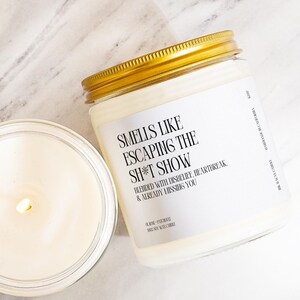 May include: A glass jar candle with a gold lid and a white label featuring black text that reads: SMELLS LIKE ESCAPING THE SH*T SHOW, BLENDED WITH DISBELIEF, HEARTBREAK, & ALREADY MISSING YOU. The candle contains white wax and a lit wick.