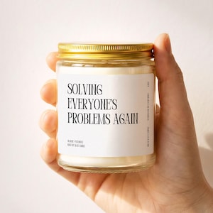 May include: A clear glass candle jar with a gold metal lid, filled with white wax. The white label features black text that reads: SOLVING EVERYONE'S PROBLEMS AGAIN, along with smaller text indicating rose, patchouli, and 100% soy wax candle.