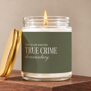 May include: A clear glass candle jar with a lit wick and a gold-colored metal lid. The label is olive green with white text that reads: SMELLS LIKE ANOTHER TRUE CRIME documentary. The candle contains white wax and is filled to the top.